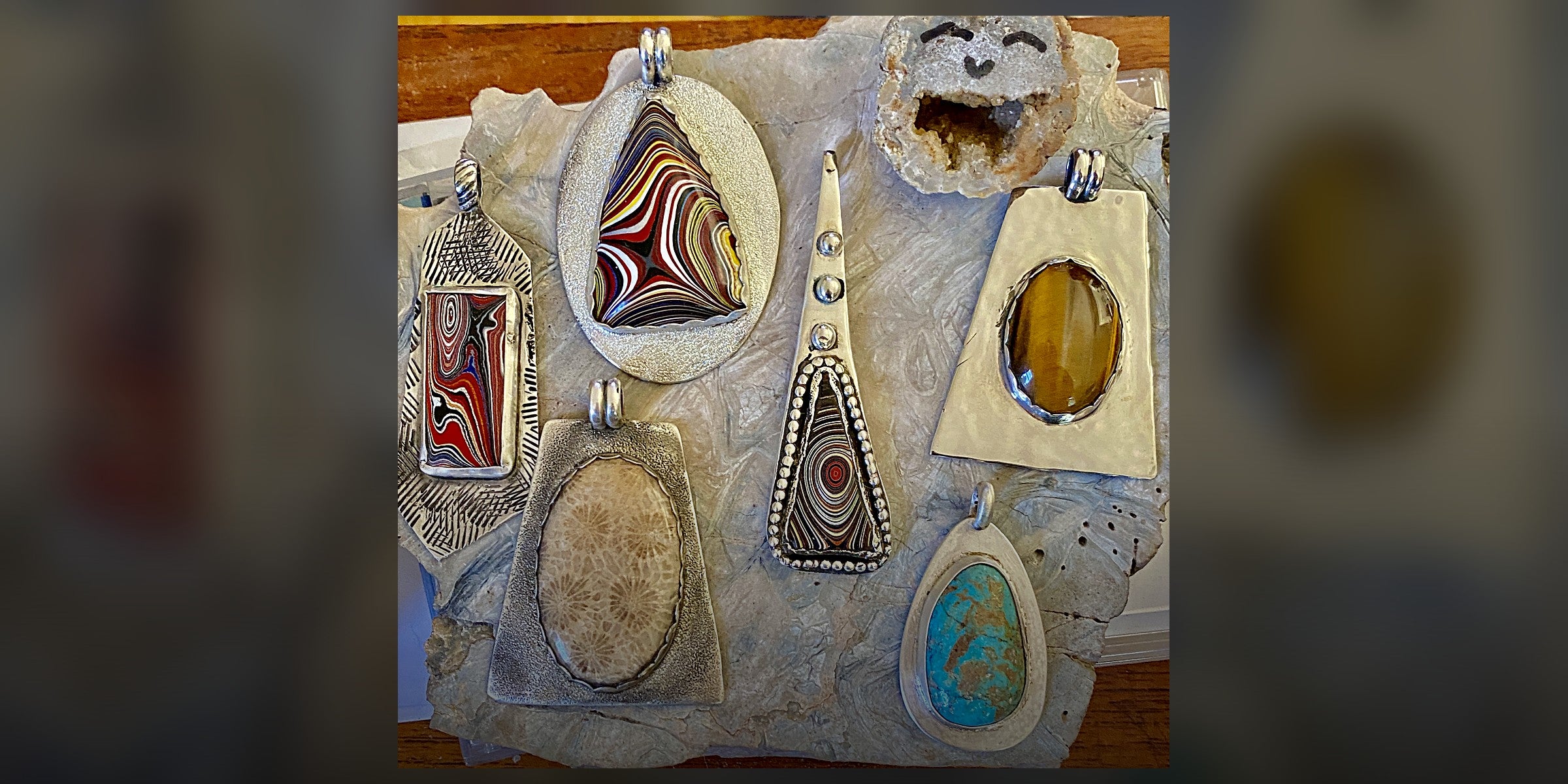Cyberzoic silver pendants — fordite, tiger's eye, turquoise, fossil coral and a druzy geode — arranged on a stone slab